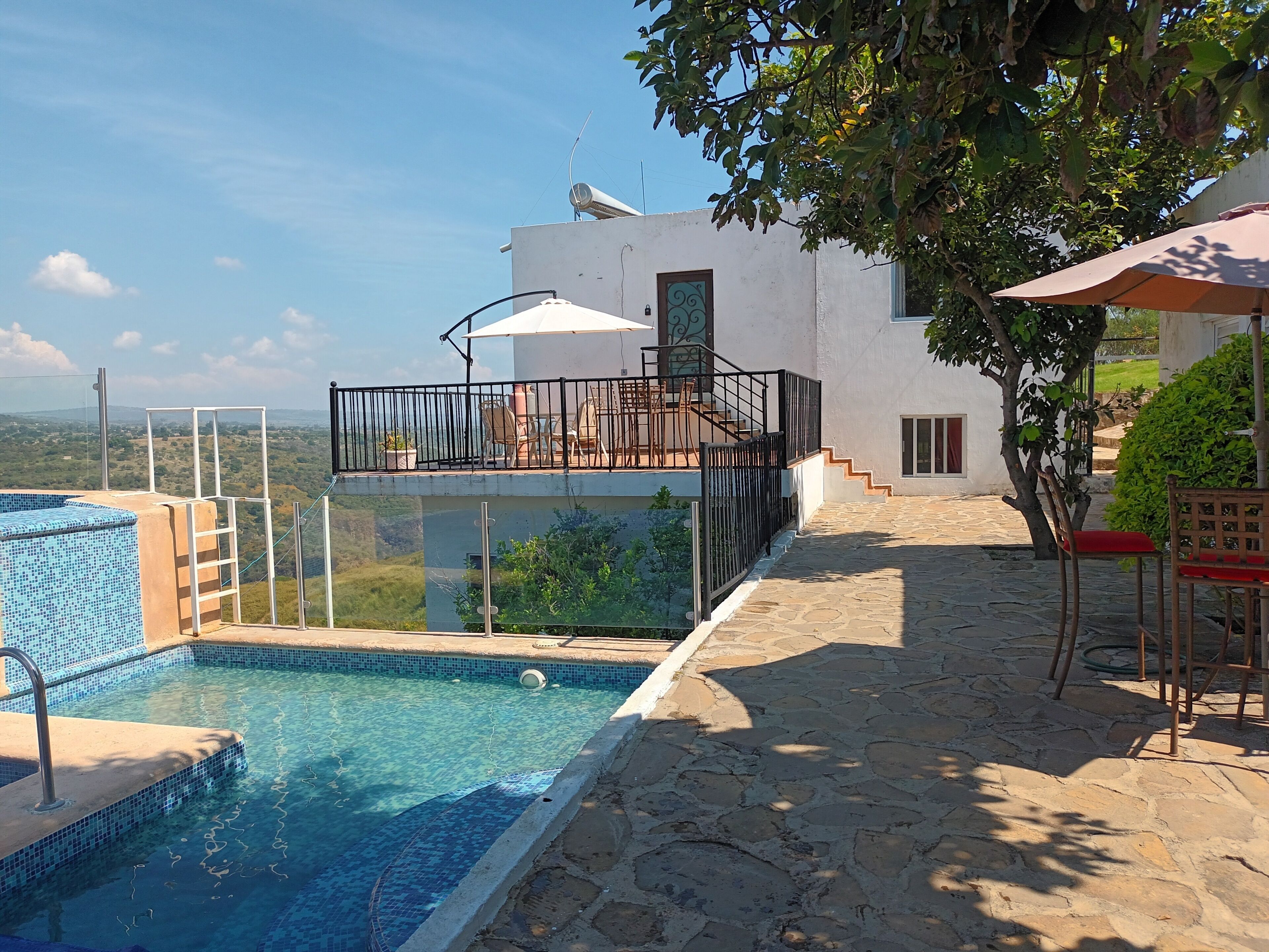 Une piscine avec un bord carrelé, un espace extérieur couvert avec des chaises, et une vue sur le paysage.