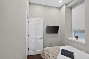 Une chambre à coucher avec un lit, un téléviseur fixé au mur, une fenêtre avec des stores et une porte.