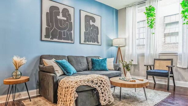 Ein modernes Wohnzimmer mit einem grauen Sofa, türkisfarbenen Kissen, einer beigen Tagesdecke und einem runden Couchtisch. An der Wand hängen zwei gerahmte Kunstwerke, eine Stehlampe und ein Sessel mit einem gewebten Rücken.