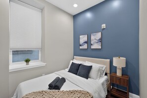 Ein Schlafzimmer mit einem Bett, einem Nachttisch, einer Lampe, einem Fenster mit Jalousien und zwei Wandbildern.
