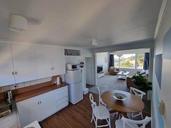 A compact kitchen with white cabinets, a microwave, and a refrigerator. There is a dining area with a wooden table and white chairs.