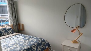 A bedroom with a bed, a bedside table with a lamp, a mirror, and a window with curtains.