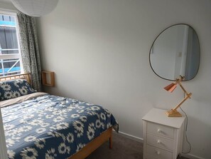 A bedroom with a bed, a bedside table with a lamp, a mirror, and a window with curtains.