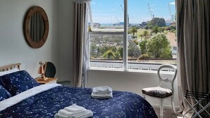 A bedroom with a bed, a chair, a window with a view of construction cranes, and a hanging light fixture. - Bay Getaway 2 Bedroom apartment (Timaru)