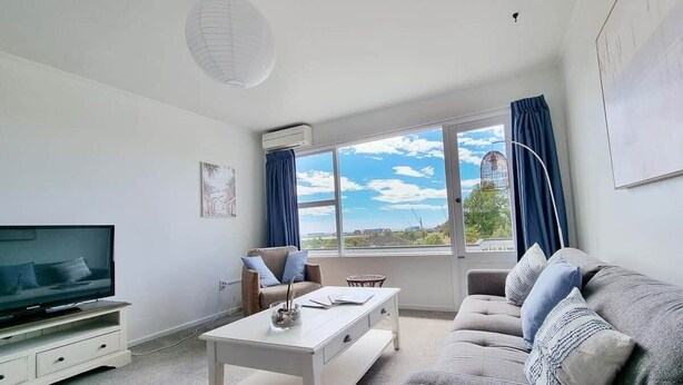 A modern living room with a flat-screen TV, a sofa, a coffee table, and a large window with a view of the sky and buildings. - Bay Getaway 2 Bedroom apartment (Timaru)