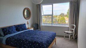 A bedroom with a bed, a chair, a window with curtains, and a mirror on the wall. - Bay Getaway 2 Bedroom apartment (Timaru)