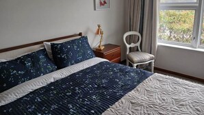 Ein Schlafzimmer mit einem Bett, einem Nachttisch, einem Sessel, einem Fenster und einem gerahmten Bild an der Wand.