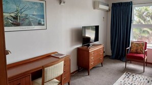 Una habitación con un escritorio de madera, un televisor en una cómoda, una silla, una ventana con cortinas y un cuadro en la pared.