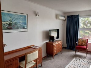 Habitació amb escriptori de fusta, televisió damunt d'una petxina, una cadira, finestra amb cortines i un quadre al mur.