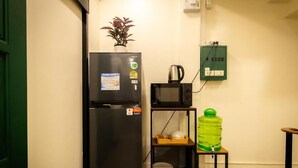 Uma cozinha compacta com geladeira preta, micro-ondas, prateleira pequena com louças, chaleira e dispenser de água verde.