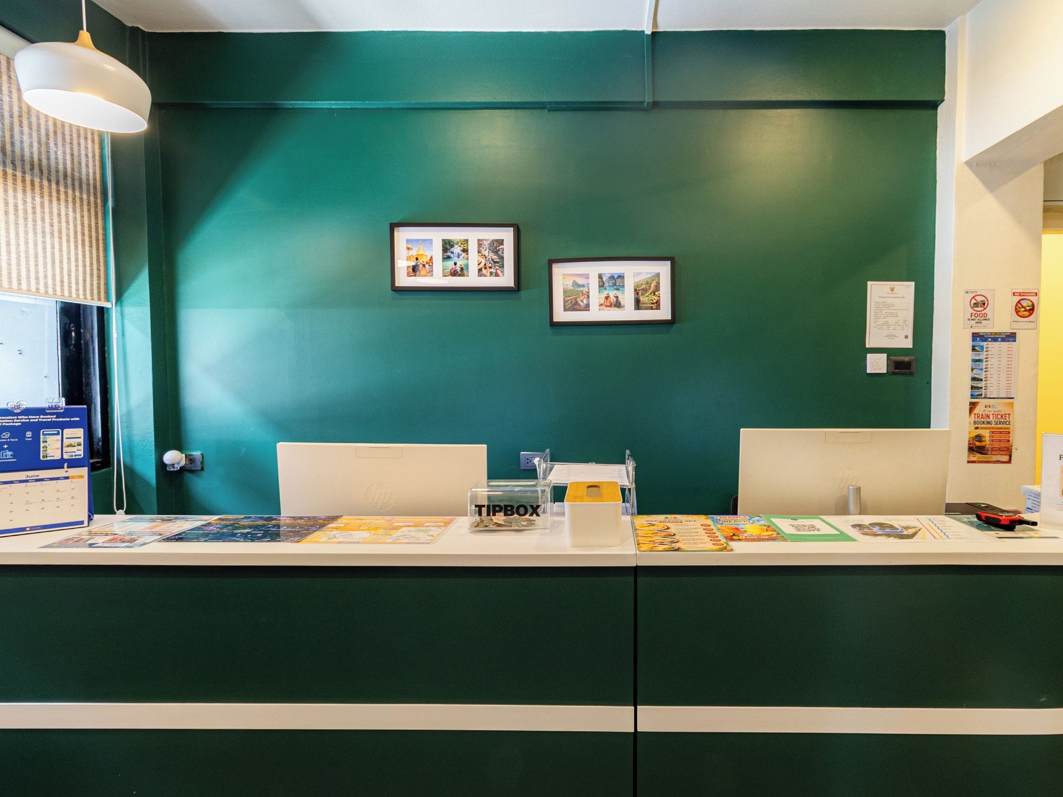 A reception desk with a green wall, framed pictures, a calendar, a tipbox, and a train ticket booking service poster.