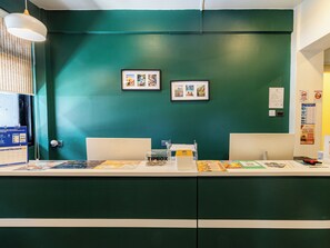 A reception desk with a green wall, framed pictures, a calendar, a tipbox, and a train ticket booking service poster. -  Social Pod Khao San Hostel (Bangkok)