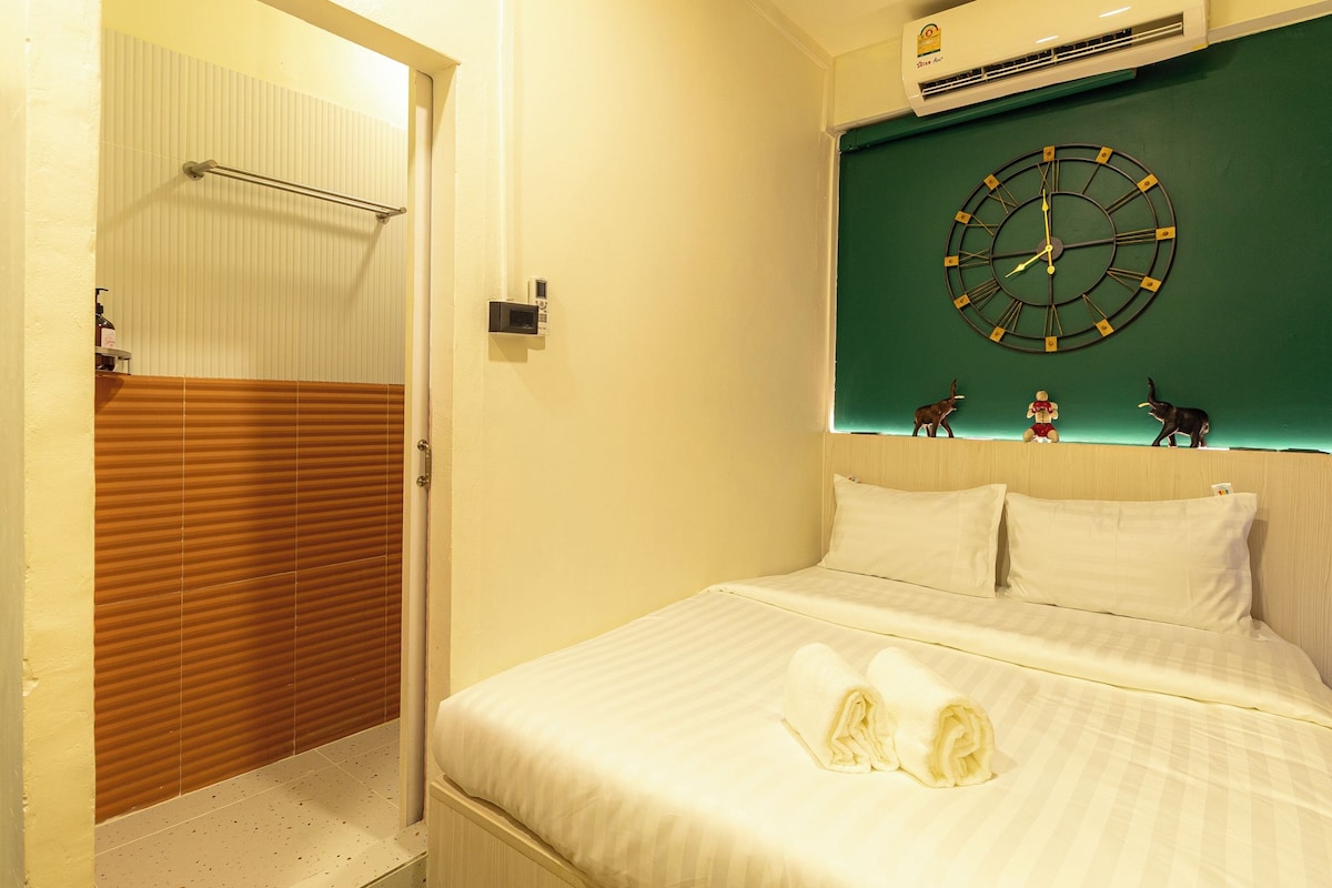 A hotel room with a bed, a wall-mounted clock, and a small shelf with figurines.