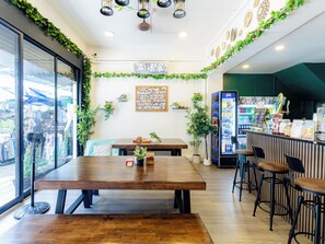 A restaurant interior with wooden tables, a bar area, and a wall decorated with greenery and photos. -  Social Pod Khao San Hostel (Bangkok)