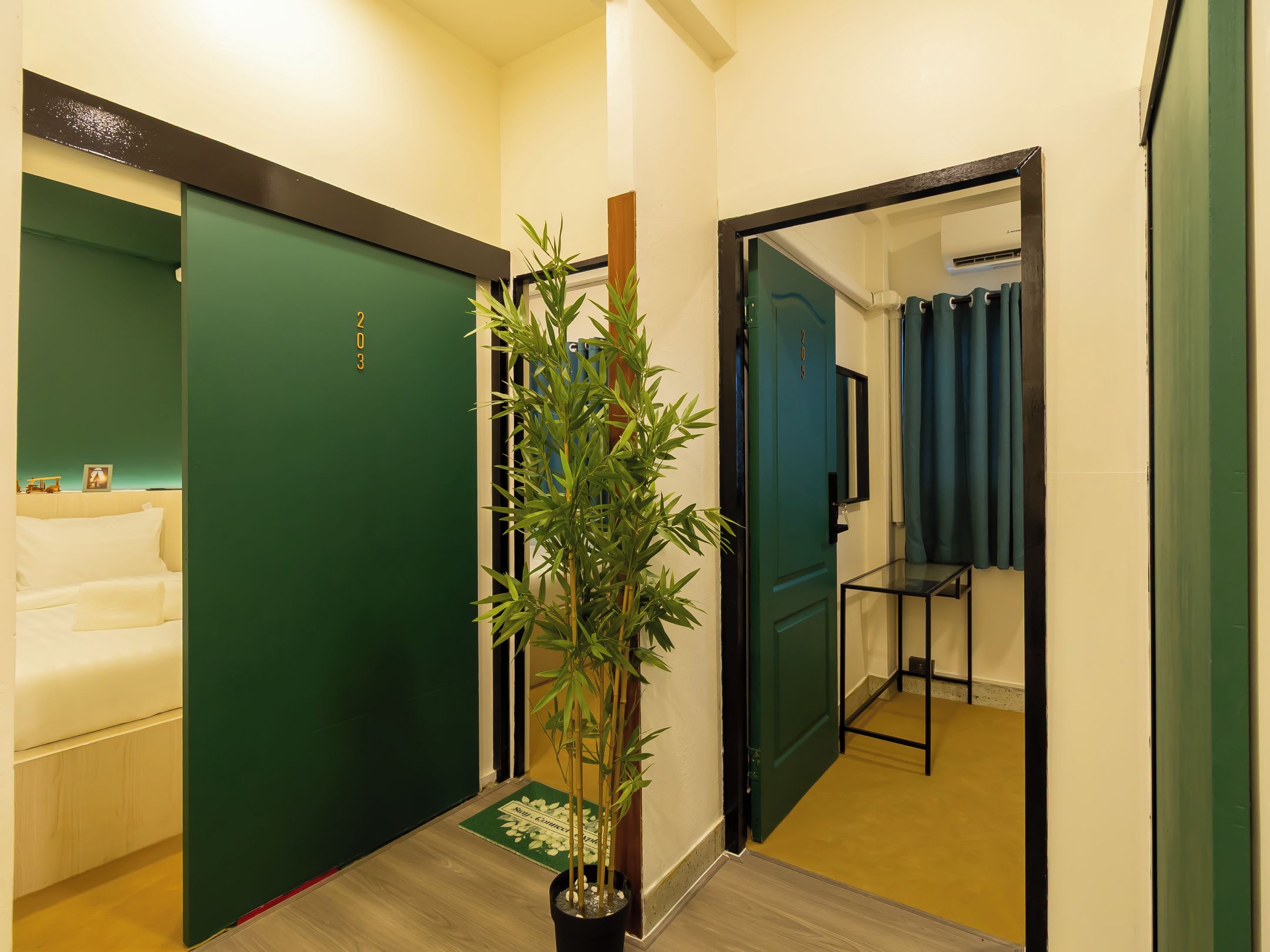 Een hotelgang met een groene deur, een kamerplant, een bed en een badkamer met een blauw douchegordijn.