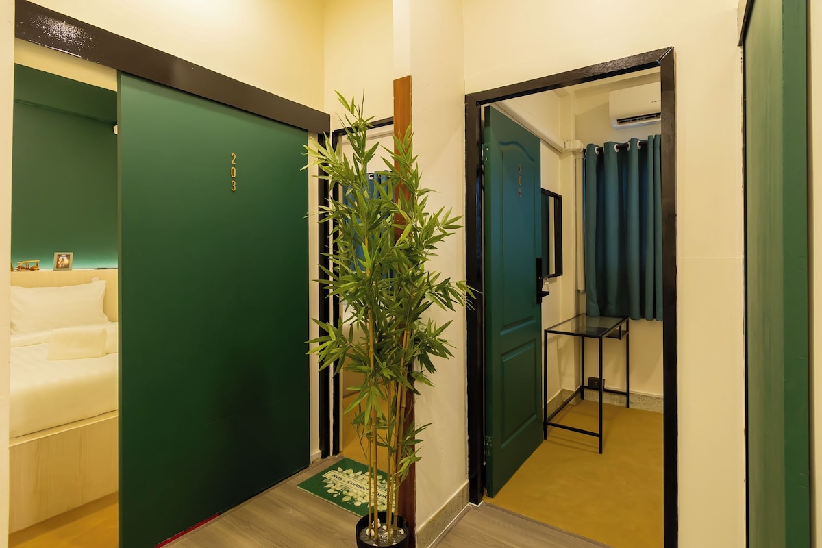 A hotel corridor with a green door, a potted plant, a bed, and a bathroom with a blue shower curtain.
