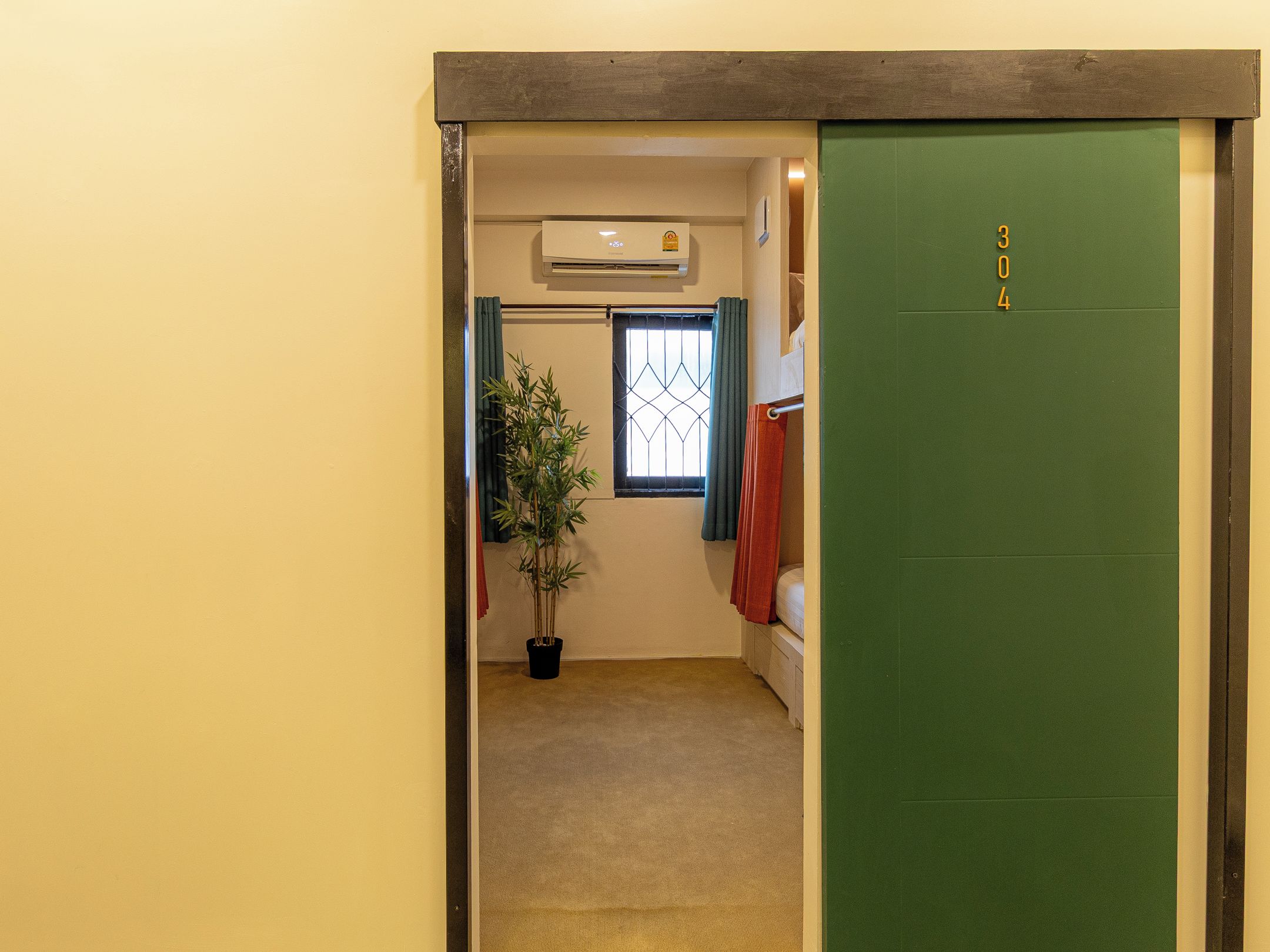Ein Flur mit einer grünen Tür, auf der '304' steht, einem Blumentopf, einem Fenster mit Vorhängen und einer Klimaanlage.