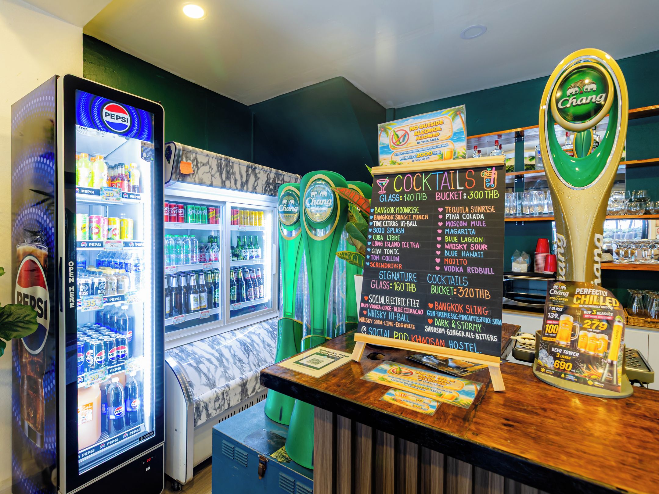 A bar area with a Pepsi refrigerator, a Chang beer tap, and a chalkboard menu listing cocktails and prices.