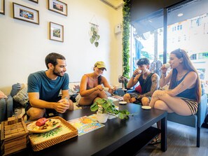 Un grupo de personas sentadas alrededor de una mesa con comida y bebidas, en una habitación con un ventanal grande y plantas de interior.