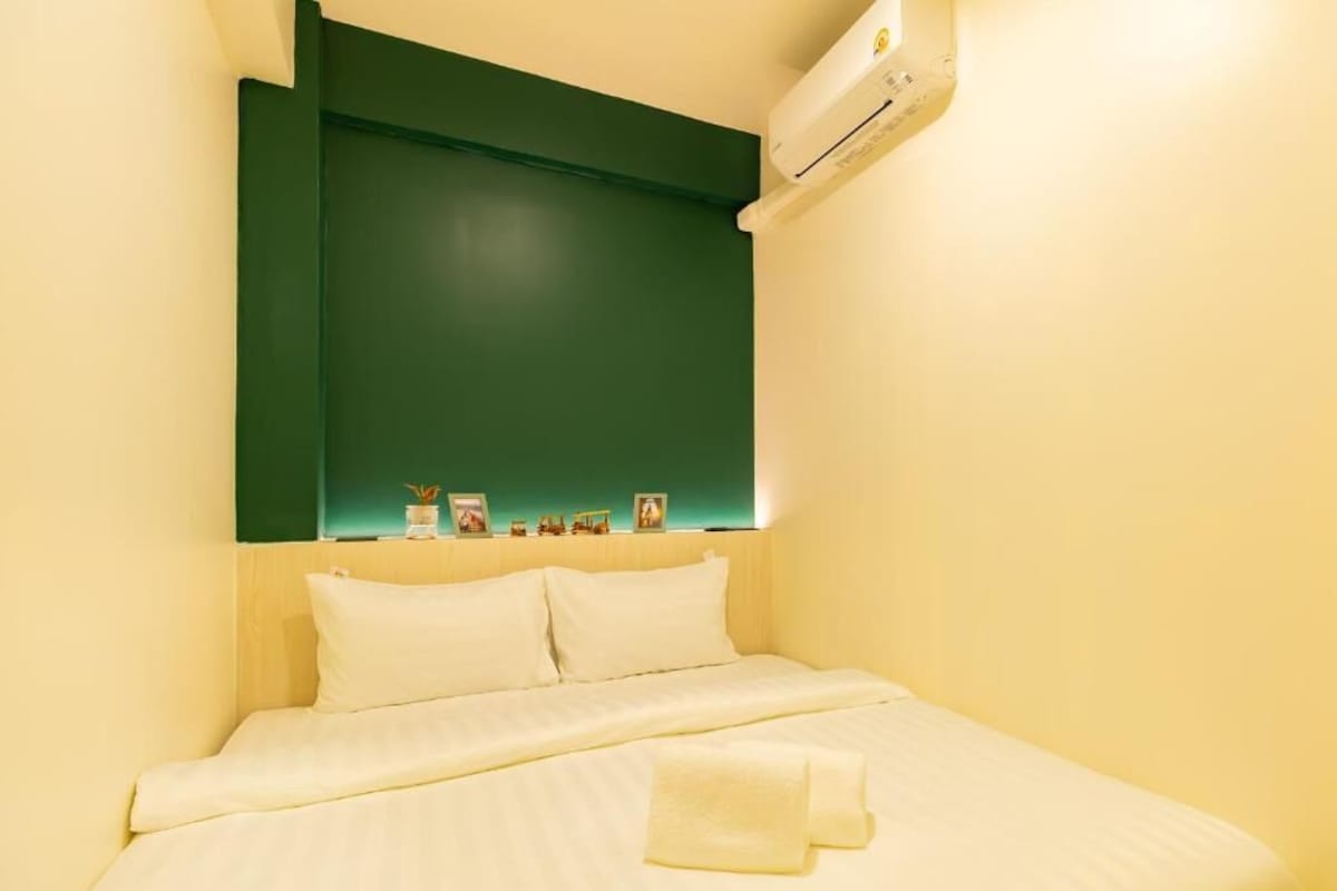 A hotel room with a bed, pillows, a bedside table with a lamp, and a wall-mounted air conditioner.