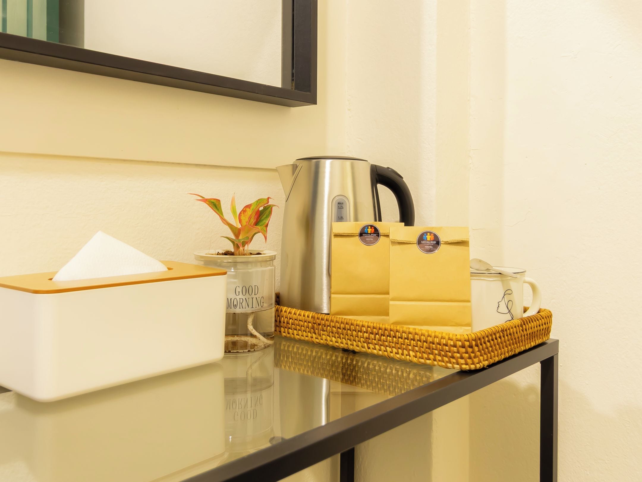 A hotel room's reception area with a coffee machine, a tissue box, a small potted plant, and a woven basket with a mug.