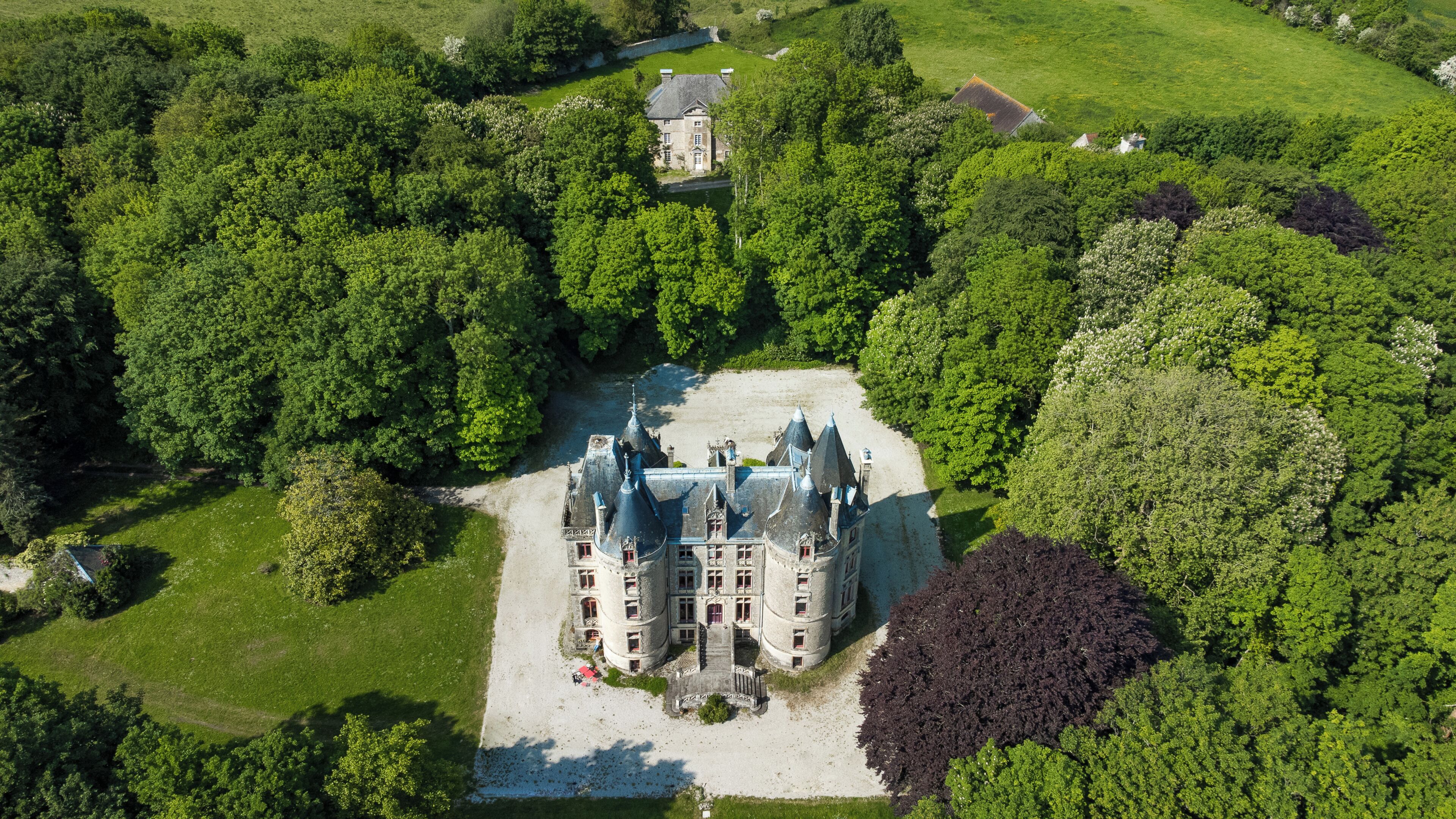 A large, historic building with multiple towers and a central courtyard, surrounded by dense greenery.