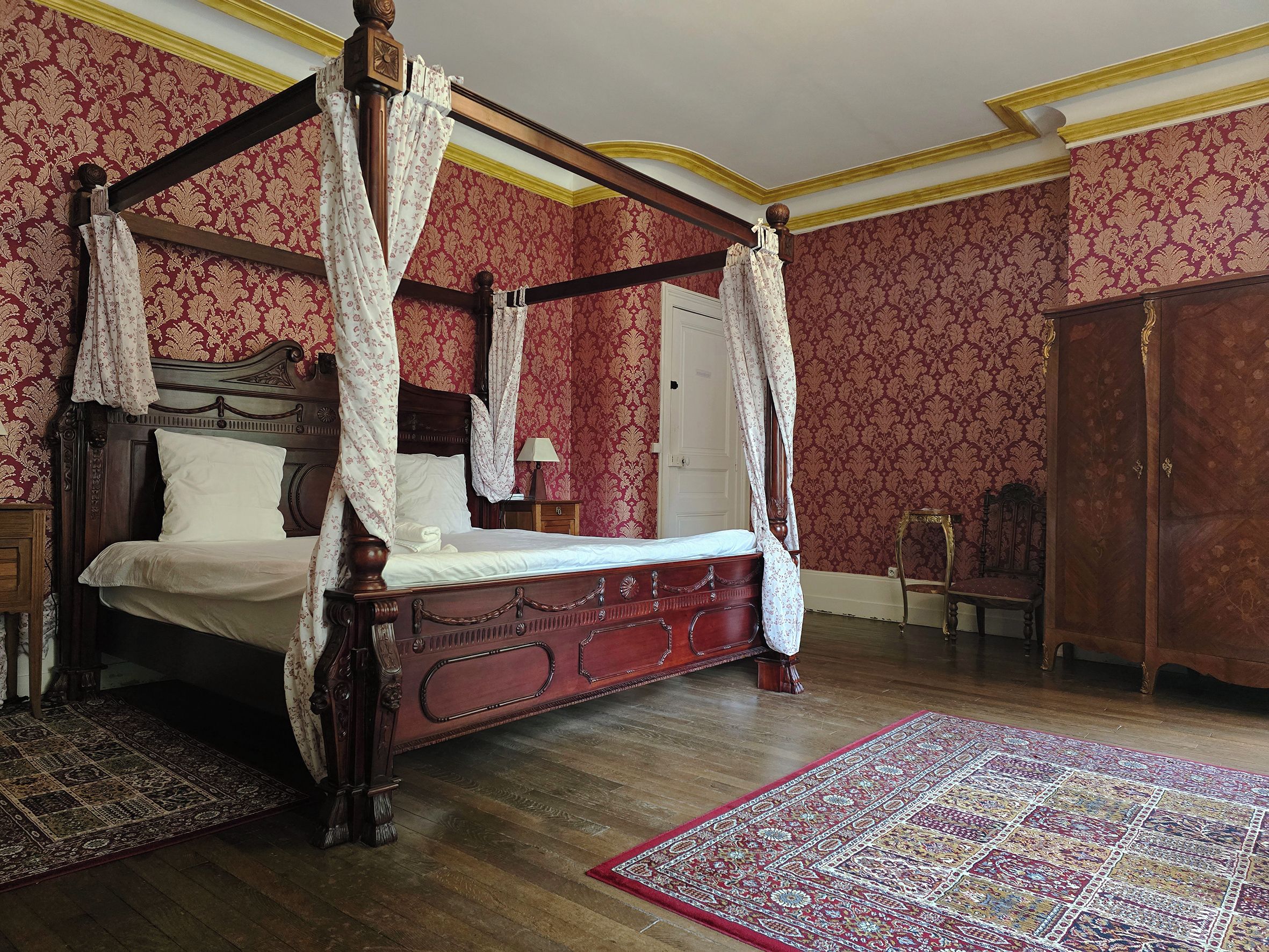 A four-poster bed with a canopy, a wooden wardrobe, and a patterned rug in a room with ornate wallpaper.