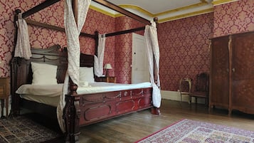 A four-poster bed with a canopy, a wooden wardrobe, and a patterned rug in a room with ornate wallpaper.