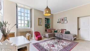 A room with a large window, a hanging light fixture, a sofa set, a patterned rug, and a door. - Château de l'Isle Marie (Picauville)