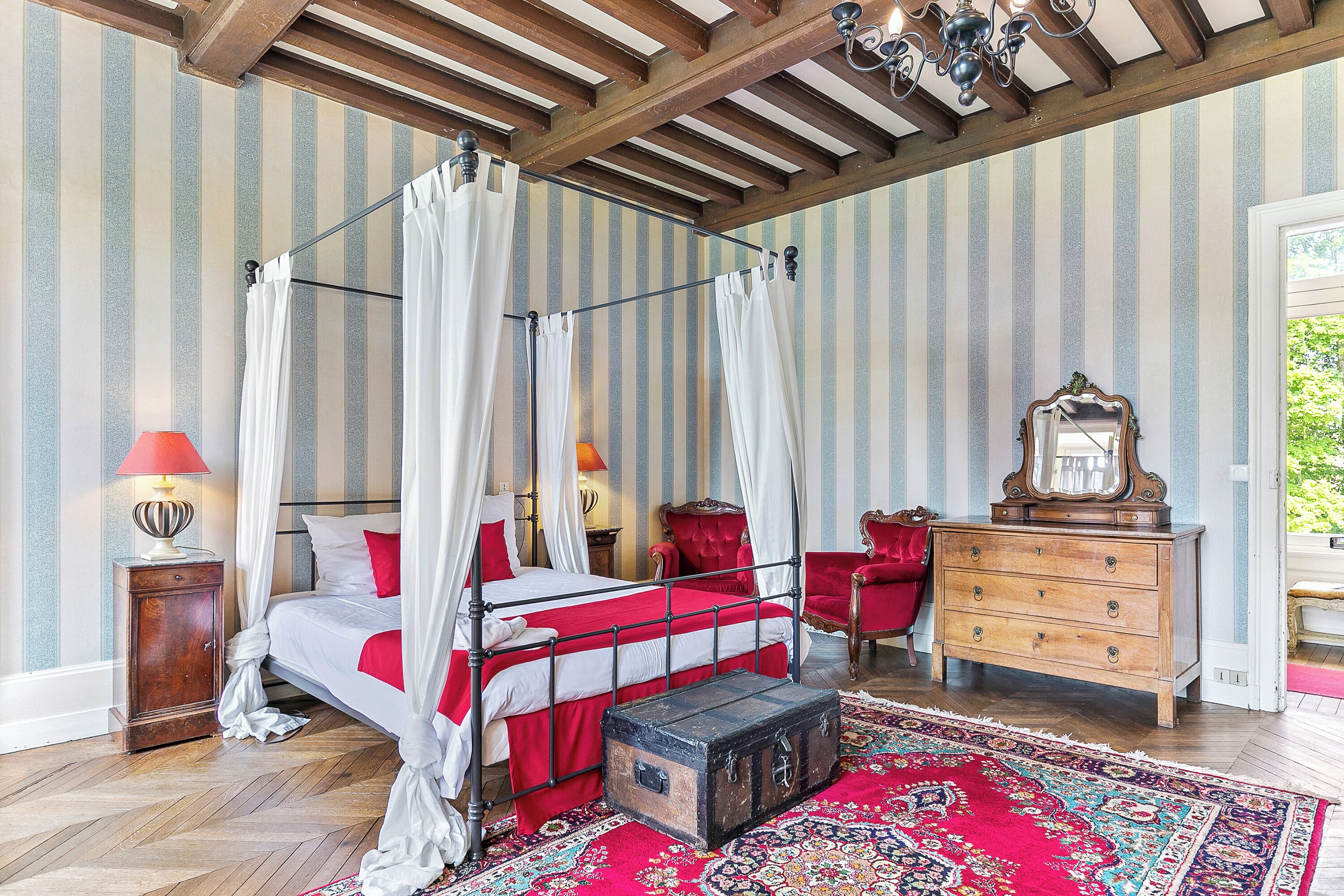 A four-poster canopy bed with a red and white bedspread, a wooden chest of drawers, a red armchair, and a patterned rug on a wooden floor.