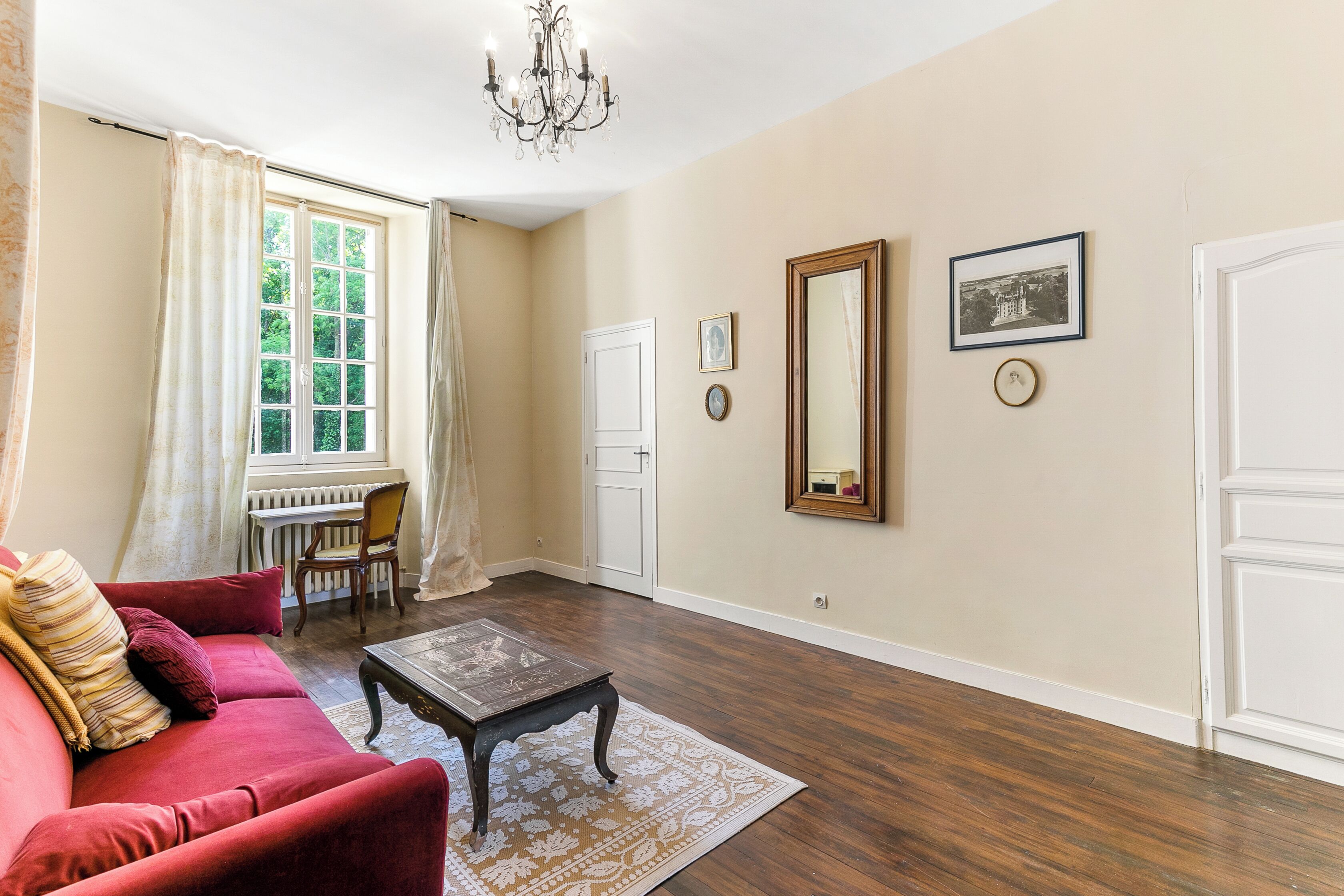 Une pièce avec un canapé rouge, une table basse en bois, un lustre, une grande fenêtre avec des rideaux et une porte.