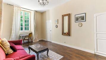 Une pièce avec un canapé rouge, une table basse en bois, un lustre, une grande fenêtre avec des rideaux et une porte.