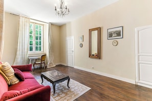 A room with a red sofa, a wooden coffee table, a chandelier, a large window with curtains, and a door.