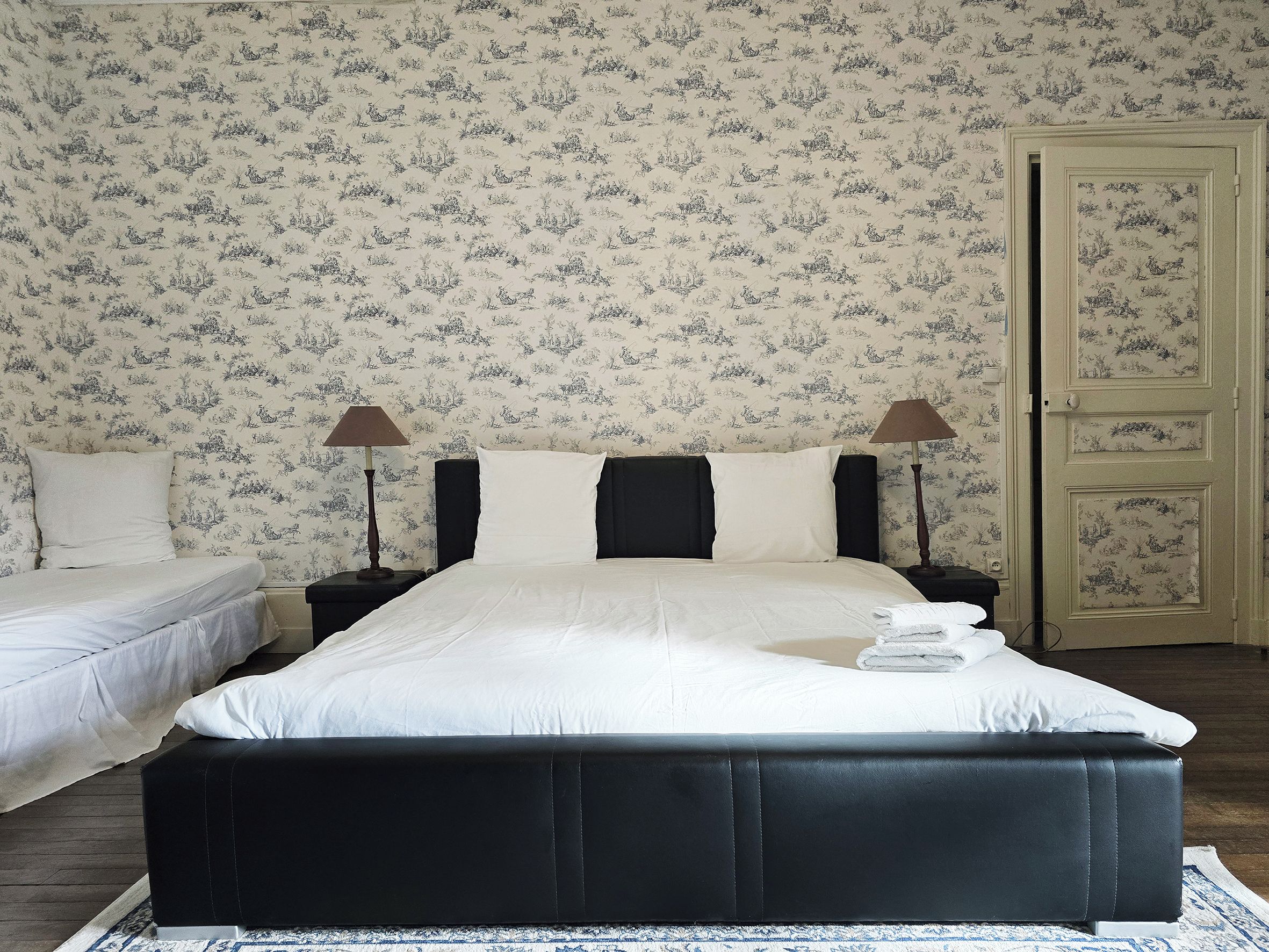 A bedroom with a large bed, two bedside tables with lamps, a door, and a patterned wallpaper.