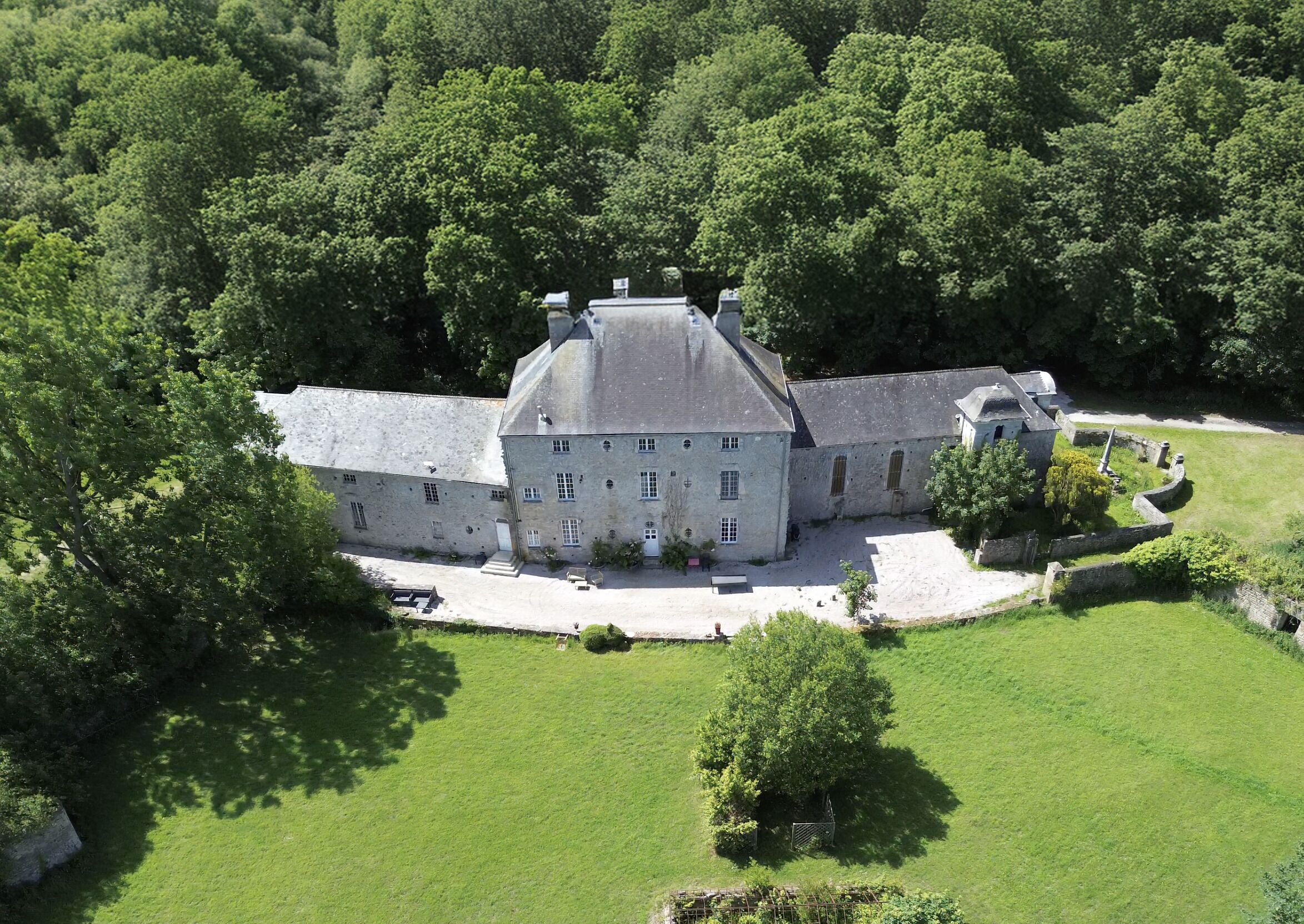 Ein großes, historisches Steinhaus mit zahlreichen Fenstern und einem zentralen Turm, umgeben von einem gepflegten Rasen und dichter Baumvegetation.