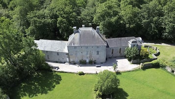 Ein großes, historisches Steinhaus mit zahlreichen Fenstern und einem zentralen Turm, umgeben von einem gepflegten Rasen und dichter Baumvegetation.
