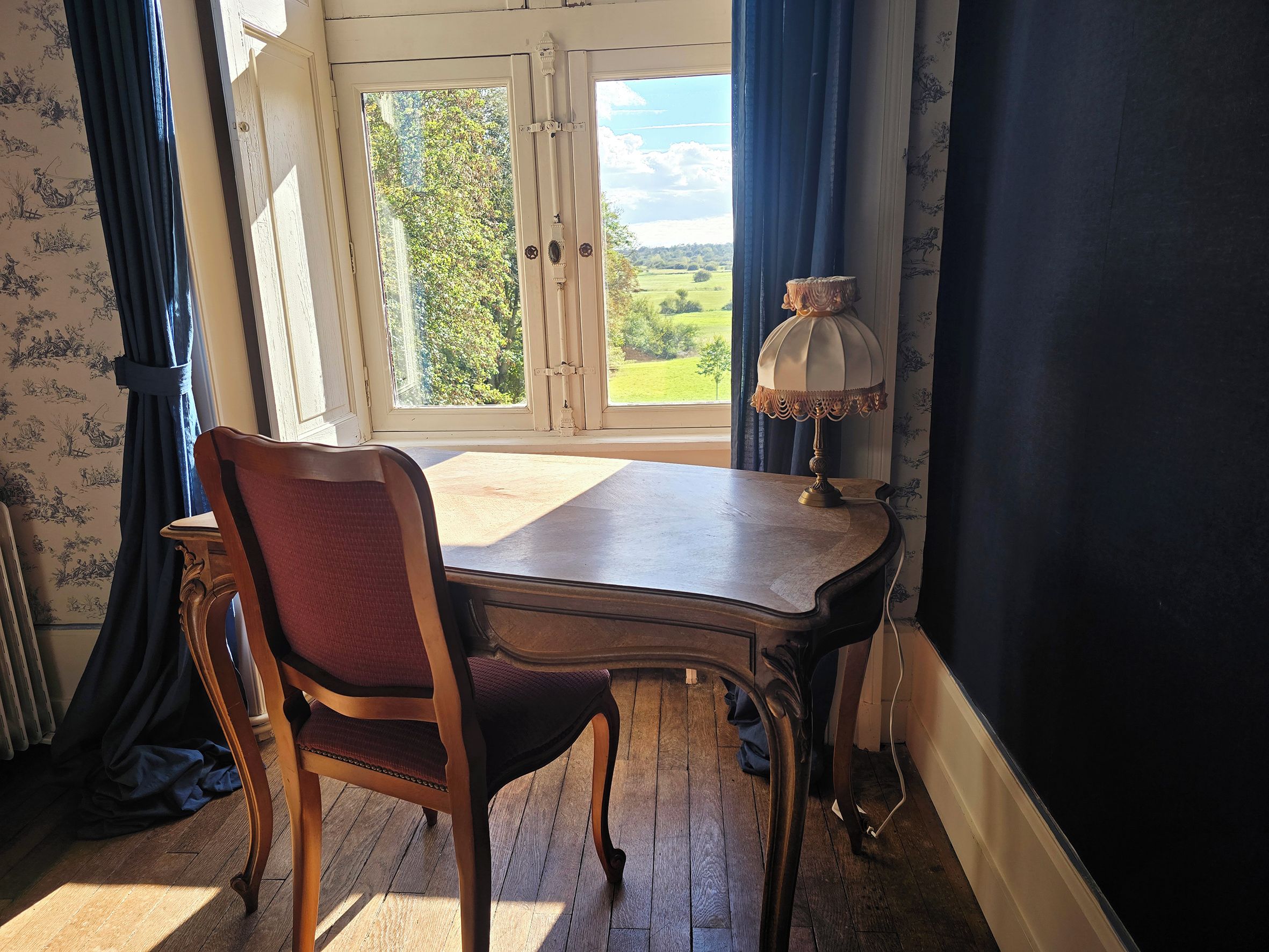 Een kamer met een houten tafel en een stoel, een raam met gordijnen en een lamp.