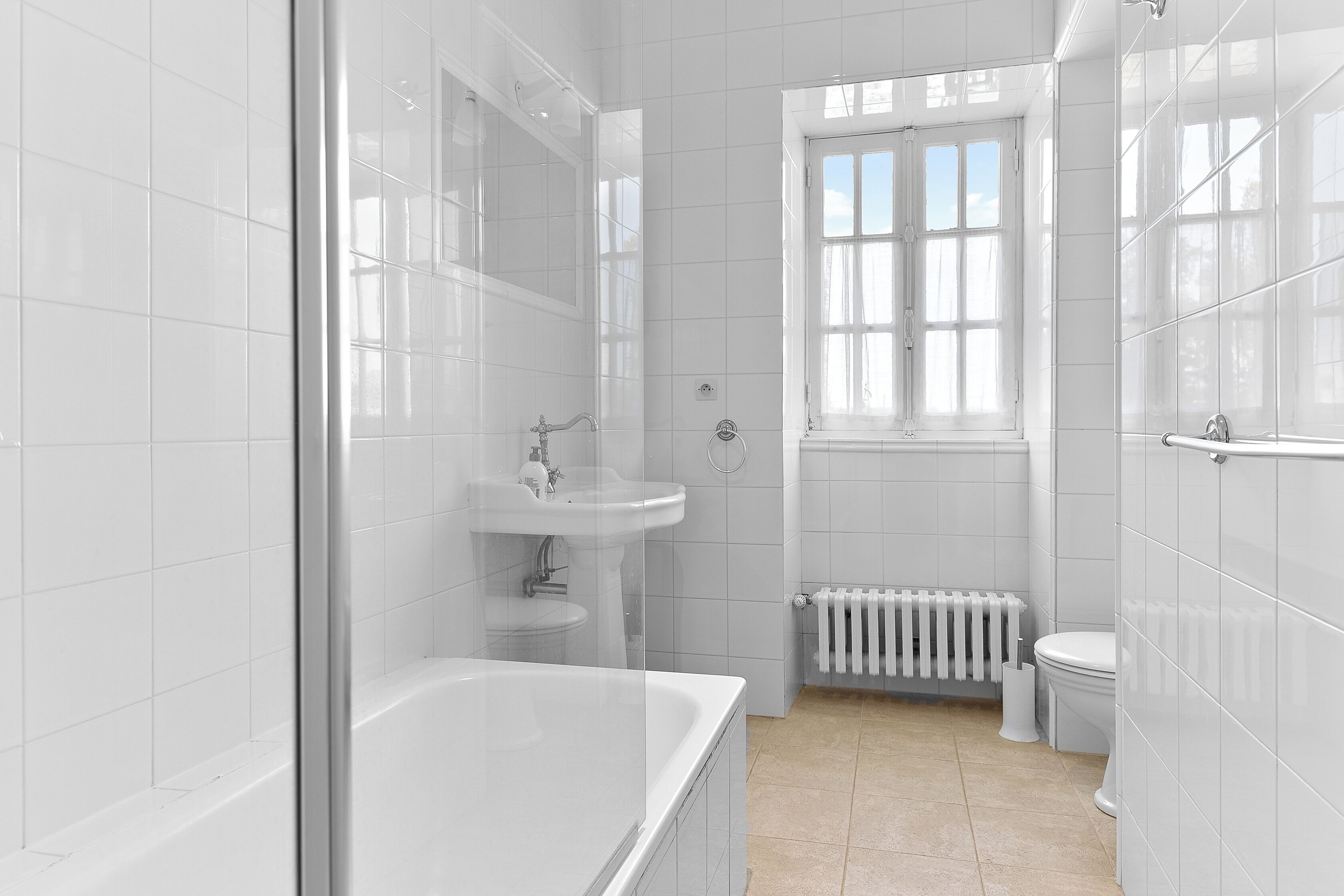 A modern bathroom with white tiles, a bathtub, a sink, a toilet, and a window.