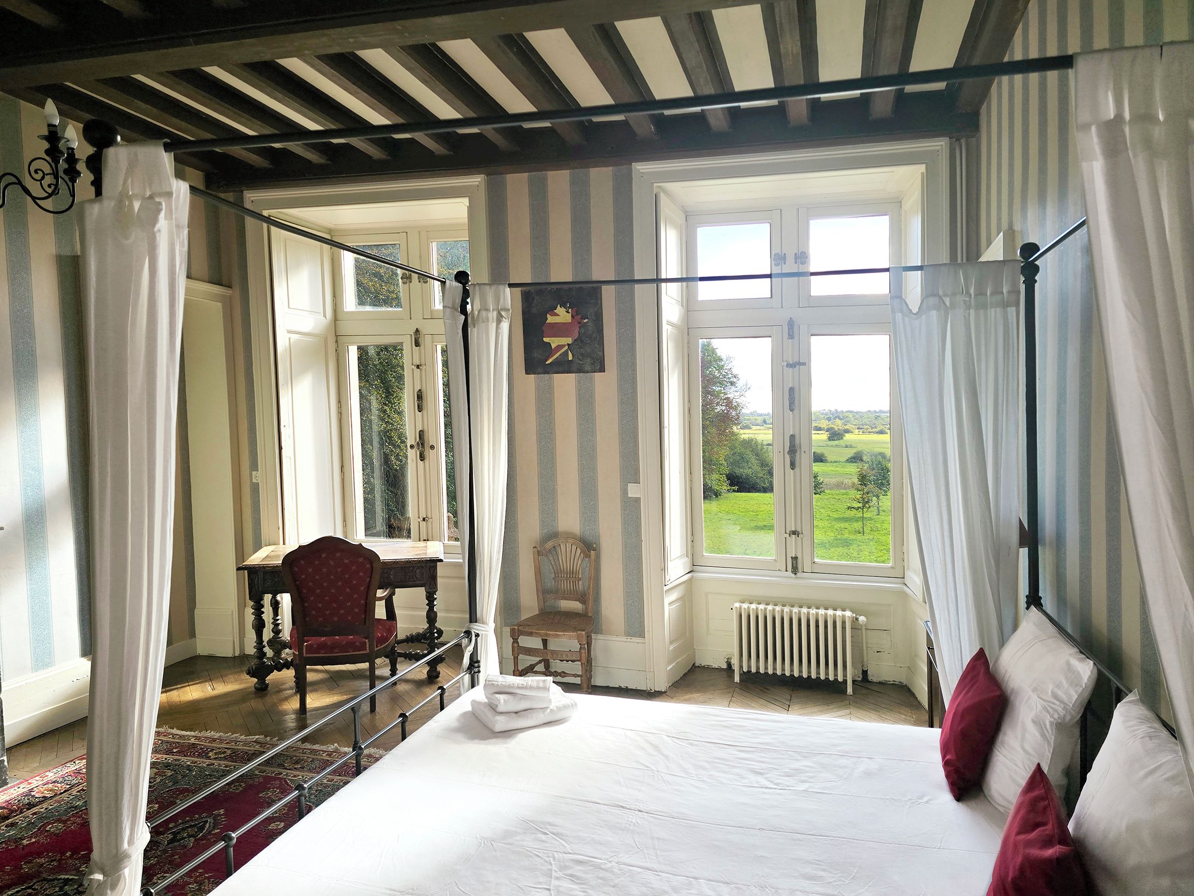 Ein Schlafzimmer mit einem Himmelbett, einem Holzstuhl, einem Heizkörper und Blick ins Freie.