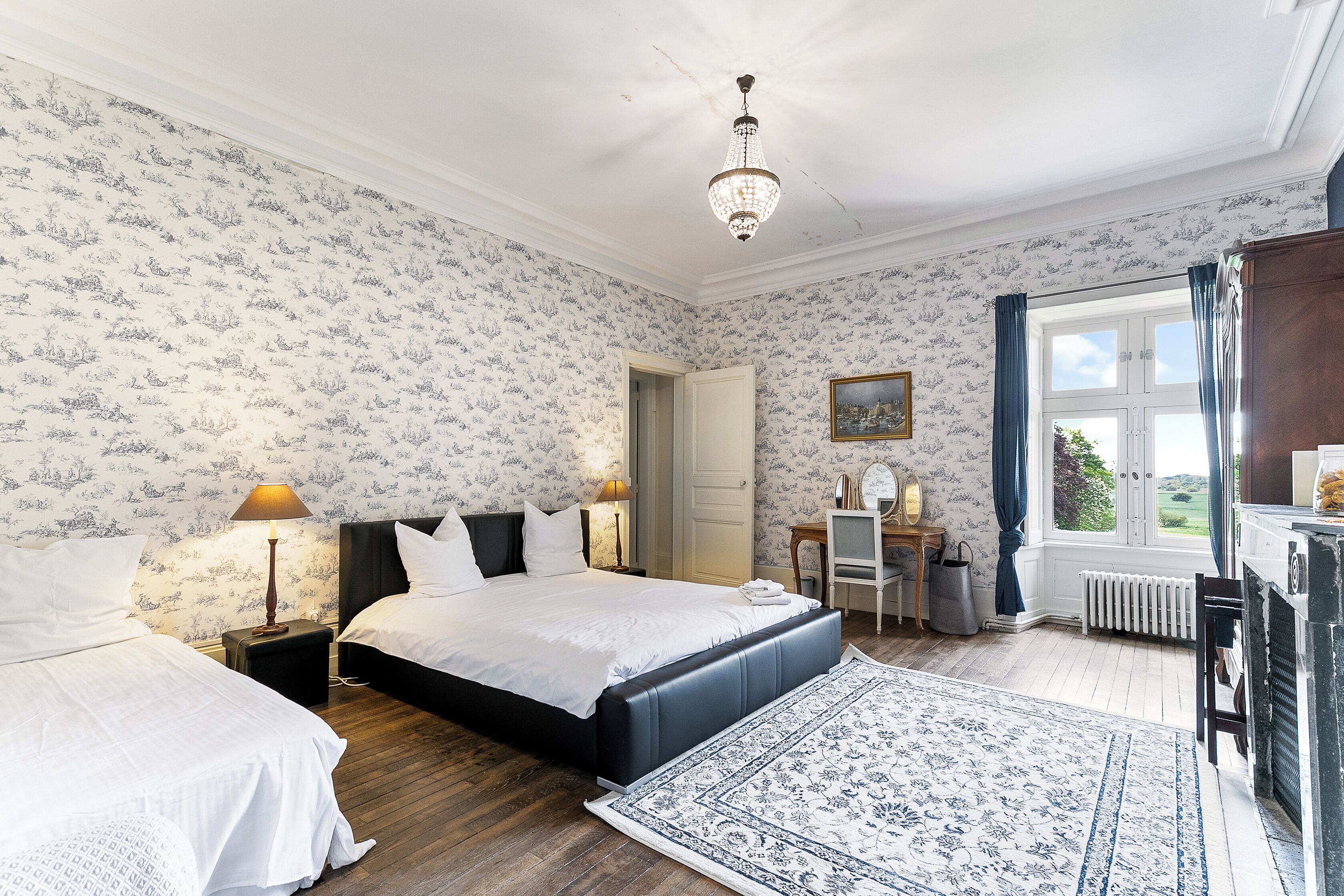 A bedroom with two beds, a desk, a radiator, and a window with curtains.