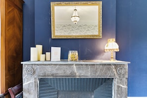 Une cheminée en marbre surmontée d’un miroir encadré, une lampe décorative et plusieurs bougies posées sur le manteau de la cheminée.