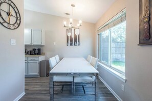 A dining area with a long table, chairs, a chandelier, a kitchen with cabinets, and a window with a view. - Family 3BR Gem with Private Pool & Game Room (Flower Mound)