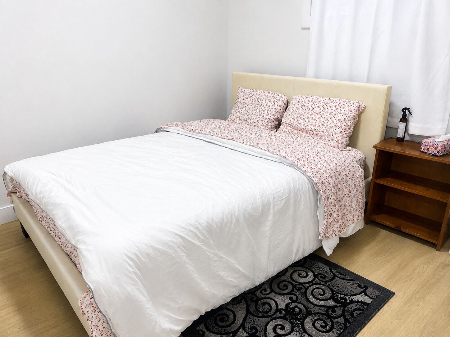 A neatly made bed with a white duvet and floral-patterned pillows, a wooden nightstand with a bottle and a tissue box, and a patterned rug on a wooden floor.