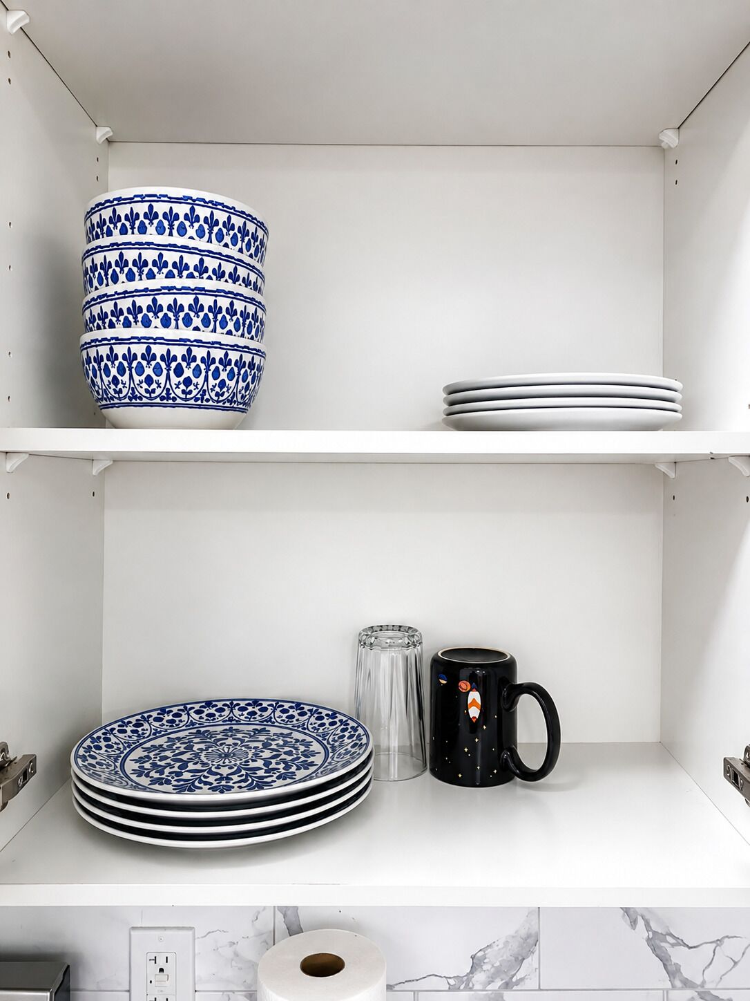 Uma prateleira na cozinha com armários brancos, uma tigela azul e branca estampada, pratos empilhados, um copo de vidro transparente e uma caneca preta com design.