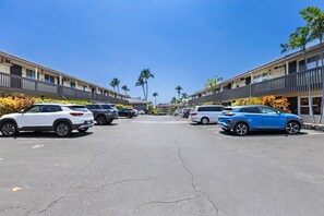 A parking lot with multiple cars parked in front of a row of buildings with balconies and palm trees. - Oceanfront 1/1 Kailua Kona Shores | Lanai + Pool (Kailua-Kona)