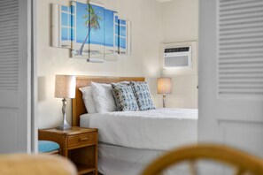 A hotel room with a bed, two bedside lamps, a nightstand, a wall-mounted air conditioner, and a beach-themed artwork above the bed. - Oceanfront 1/1 Kailua Kona Shores | Lanai + Pool (Kailua-Kona)