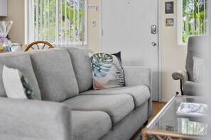 Ein modernes Wohnzimmer mit einem grauen Sofa, einem Glastisch und Blick auf Grün durch das Fenster.