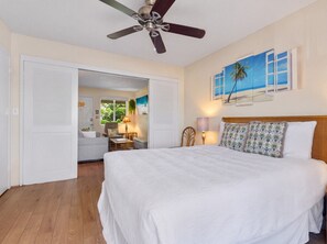Ein Hotelzimmer mit einem Bett, einem Deckenventilator, einem Schreibtisch mit Stuhl, einer Lampe und einem Strandbild.