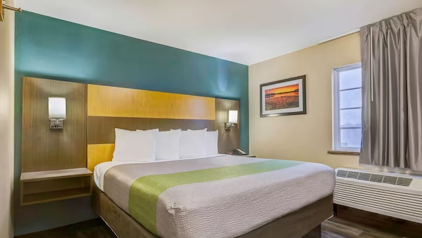 A hotel room with a large bed, bedside tables, a wall-mounted lamp, a painting, and a window with curtains. - 1 King Bed Unit w/ Kitchenette & Free Parking | Oak Park Mall - 2.4 miles (Johnson County)