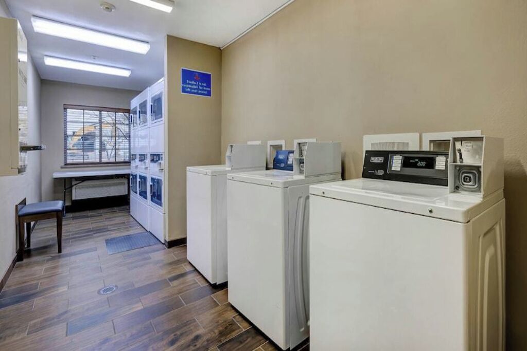 A laundry room with three washing machines, a bench, and a window with blinds.